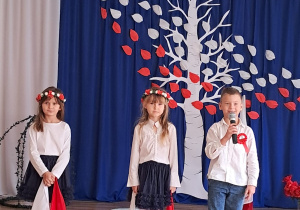 Dwie dziewczynki w wiankach z biało-czerwonych kwiatów trzymają chustki w barwach narodowych i chłopiec, który mówi do mikrofonu.