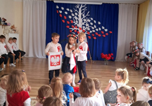 Chłopiec trzyma godło Polski, dziewczynka mówi do mikrofonu obok niej stoi drugi chłopiec.