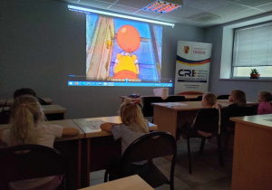 Dzieci siedząc w szkolnych ławeczkach oglądają film edukacyjny o Kubusiu Puchatku.