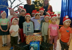 Pani Dyrektor, p. Grażynka w mikołajkowych czapkach, oraz grupa dzieci z naszego przedszkola wraz z paczkami przygotowanymi na akcję charytatywną, pozują na tle choinki.