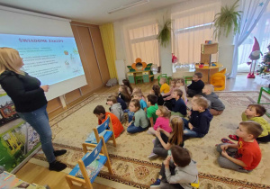 Dzieci siedząc na dywanie z uwagą słuchają wygłaszanej przez pania z PGK-u pogadanki na temat prawidłowej segregacji odpadów. Oglądaja również tematyczne obrazki.