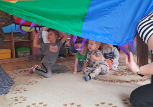 Dzieci siedzą na dywanie pod chustą animacyjną trzymaną przez dorosłych.