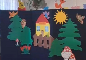 Kadr z teatrzyku sylwet pod tytułem Leśne przedszkole. Dekoracja to: drzewa, budynek przedszkola, ptaki, słońce, muchomory oraz sylwety zajączka i liska.