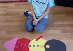Chłopiec ułożył na podłodze bałwana z kolorowych papierowych figur geometrycznych.
