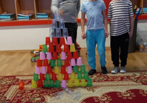 Trzech chłopców stoi za bardzo dużą budowlą, którą ustawili z kolorowych kubków jednorazowych.