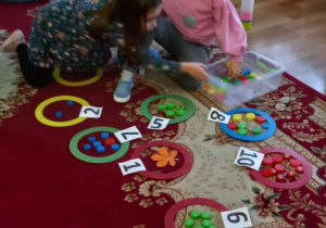 Dwie dziewczynki układają elementy w kołach oznaczonych cyfrą. Tworzą zbiory.