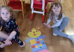 Dwie dziewczynki siedzą na podłodze, między nimi sylweta lalki z kolorowych, papierowych figur geometrycznych, którą same ułożyły.