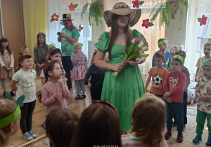 Przedszkolaki a wśród nich kobieta przebrana za Wiosnę w zielonej sukni i słomkowym kapeluszu.