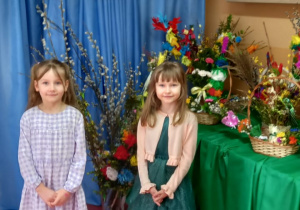 Dwie dziewczynki nad nimi napis Palmy Wielkanocne, za nimi bazie wierzbowe w wazonie i koszyczki z palmami i ozdobami wielkanocnymi.