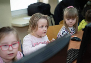 Trzy dziewczynki siedzą przed monitorami komuterów.