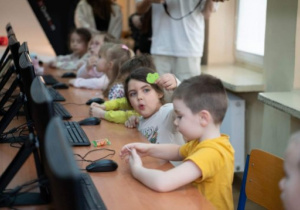 Dzieci siedzą przy pulpicie z komputerami.