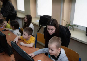 Dzieci siedzą przed monitorami, wykonują jakieś zadanie, dziewczyna stoi za nimi nadzoruje ich pracę.