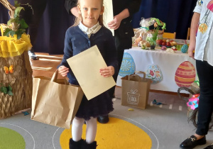 Dziewczynka trzyma dyplom i papierową torbę z nagrodą za udział w konkursie, za nią dwie kobiety organizatorki konkursu.