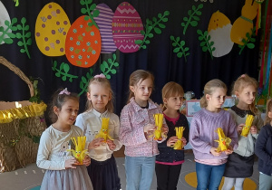 Sześć dziewczynek stoi na tle granatowej dekoracji z napisem Wielkanocna Pisanka, pod napisem duże, kolorowe, wycięte z papieru pisanki. Dzieci trzymają żółte zabawki-kurczaczki.