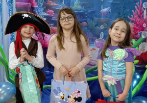Natan w przebraniu pirata, Emilka w różowej sukience i Kornelka w stroju syrenki. Dzieci trzymają torby z nagrodami, stoją na tle dekoracji przedstawiającej morskie głębiny.