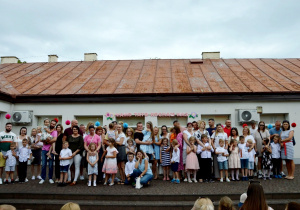 Pamiątkowe zdjęcie dzieci z grupy Jagódki z rodzicami na tarasie przedszkola. Z tyłu napis Mamo, Tato kocham Was.