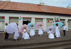 Sześć dziewczynek, ze starszaków, w białych długich sukniach trzymają rozłożone parasole i tańczą dla rodziców.