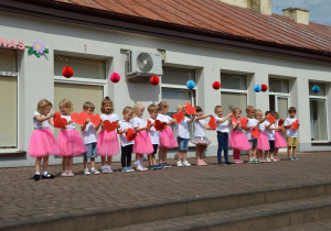 Dzieci z grupy Promyczki trzymają serca z napisem Kochamy Was, występują dla rodziców. Dziewczynki w białych koszulkach i różowych spódniczkach.