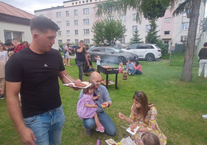 Mężczyzna trzyma tackę z kiełbaską z grilla, rodzina zjada potrawy piknikowwe. W tle mężczyzna piecze na grillu kiełbaski.