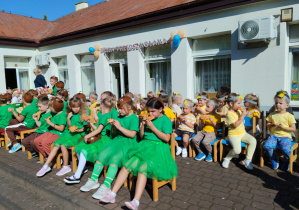 Dzieci z grupy Żuczki i Słoneczka siedzą na krzesełkach na patio przed przedszkolem i jedzą pizzę.