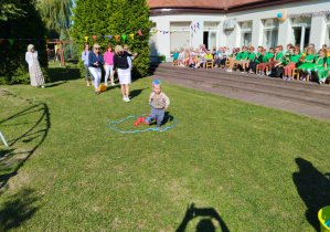 Chłopiec z grupy Jagódki biegnie z niebieską piłeczką. Za nim nauczyciele podczas konkurencji przenoszenia piłeczek.