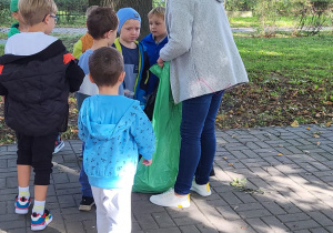 Kobieta trzyma zielony worek na odpady, a dzieci wrzucają do niego uzbierane w parku śmieci.