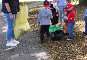 Kobieta trzyma żółty worek. Dzieci wrzucają odpady zebrane na trawniku do zielonego worka.