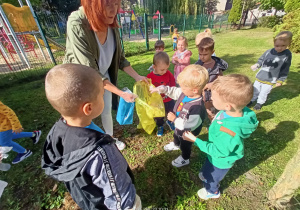 Kobieta trzyma żółty worek dzieci wrzucają do niego plastikowe śmieci z ogrodu przedszkola.