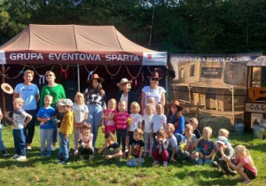 Grupa przedszkolaków stoi na tle dekoracji będącej atrapą Dzikiego Zachodu. Pozują z nauczycielkami i animatorką w westernowych kapeluszach.