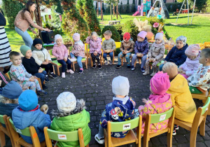 Grupa najmłodsza 3-latków, siedząc na zielonych krzesełkach, w kółeczku na tarasie, prezentuje swoje ziemniaki konkursowe, trzymając je na kolankach.