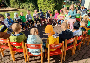 Grupa 6-latków, siedząc na czerwonych krzesełkach, w kółeczku na tarasie, prezentuje swoje ziemniaki konkursowe, trzymając je wysoko nad głowami.