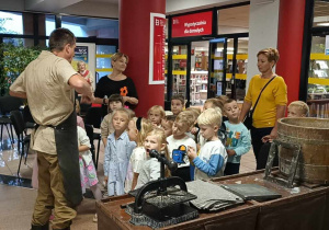 Dzieci i panie słuchają z ciekawością opowiadań pana prowadzącego warsztaty, stojąc przy stole z narzędziami do produkcji papieru..