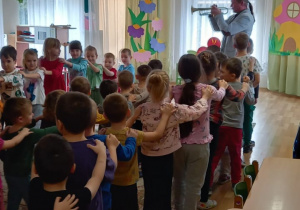 Przedszkolaki stoją jeden za drugim trzymając ręce na ramionach dziecka z przodu, utworzyły pociąg. Muzyk gra na trąbce.