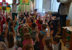 Dzieci siedzą na dywanie i klaszczą nad głowami, pośrodku stoi chłopiec i też klaszcze nad głową. Obok muzyk gra na trąbce.