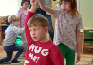 Chłopiec klęczy na jednym kolanie ma uniesioną rękę, którą trzyma dziewczynka tańcząca wokół niego. Z tyłu stoi mężczyzna trzyma w ręce trąbkę, drugą rękę ma uniesioną.