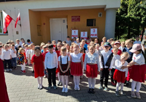Dzieci stoja przed przedszkolem, udekorowanym tablicami upamiętniającymi święto 1,2 i 3-maja.