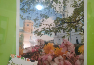 Bukiet kolorowych kwiatów nad nim widać wieżę ratuszową muzeum w Radomsku i biały kościół.