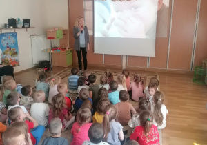 Dzieci siedzą na dywanie i oglądają prezentację multimedialną, którą prowadzi pani dietetyk.