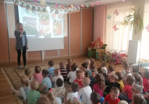 zieci siedzą na dywanie i oglądają prezentację multimedialną, którą prowadzi pani dietetyk.