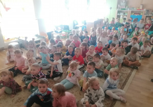 Dzieci siedzą na dywanie w sali przedszkolnej, niektóre z nich podniosły rękę.