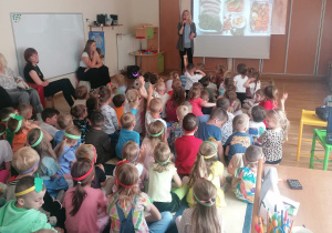 zieci siedzą na dywanie i oglądają prezentację multimedialną, którą prowadzi pani dietetyk.