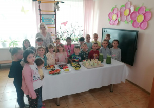 Dzieci z nauczycielką stoją wokół stołu z owocowymi przekąskami i sokiem owocowym.