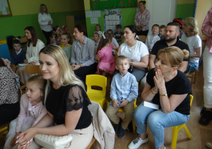 Dzieci i ich opiekunowie siedzą na widowni w sali przedszkolnej.