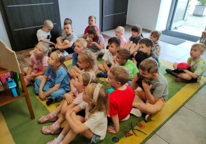 Grupa dzieci przedszkolnych siedzi w sali biblioteki na dywanie i słucha bajkę.