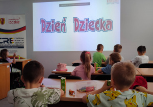 Dzieci siedzą przy stolikach przed nimi wyświetlony kolorowy napis Dzień Dziecka