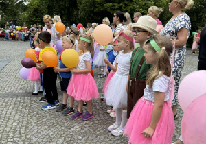 Dzieci kolorowo ubrane trzymają balony za nimi stoją opiekunowie.
