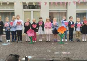 Dzieci stoją na patio przedszkola, trzymają sylwety czerwonych serc. Za nimi napis Mama i Tata najpiękniejsze słowa świata.