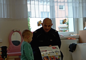 Pan policjant trzyma planszę z numerami alarmowymi, obok stoi chłopiec i uważnie przygląda się planszy.