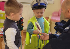 Chłopiec w żółtej, odblaskowej kamizelce trzyma lizak policyjny, a na głowie ma czapkę policyjną. Pan policjant coś mówi do niego i jego kolegi.