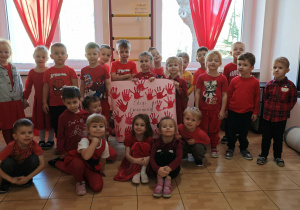 Dzieci z grup: Promyczki i Motylki, ubrane w czerwone bluzki, trzymają plakat, na którym są czerwone odciski dłoni wycięte z papieru i napis Stop przemocy!